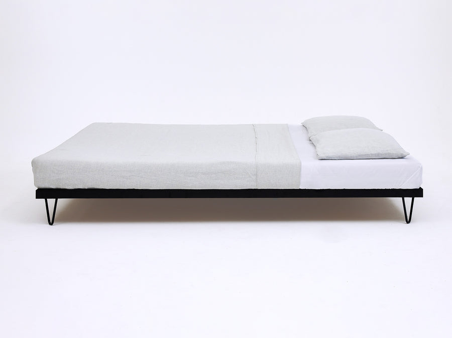 Seitenansicht Kitanda Bettgestell aus Stahl mit filigranen V-förmigen Füßen, weißem Bettlaken und hellgrauer Bettwäsche vor weißem Hintergrund
