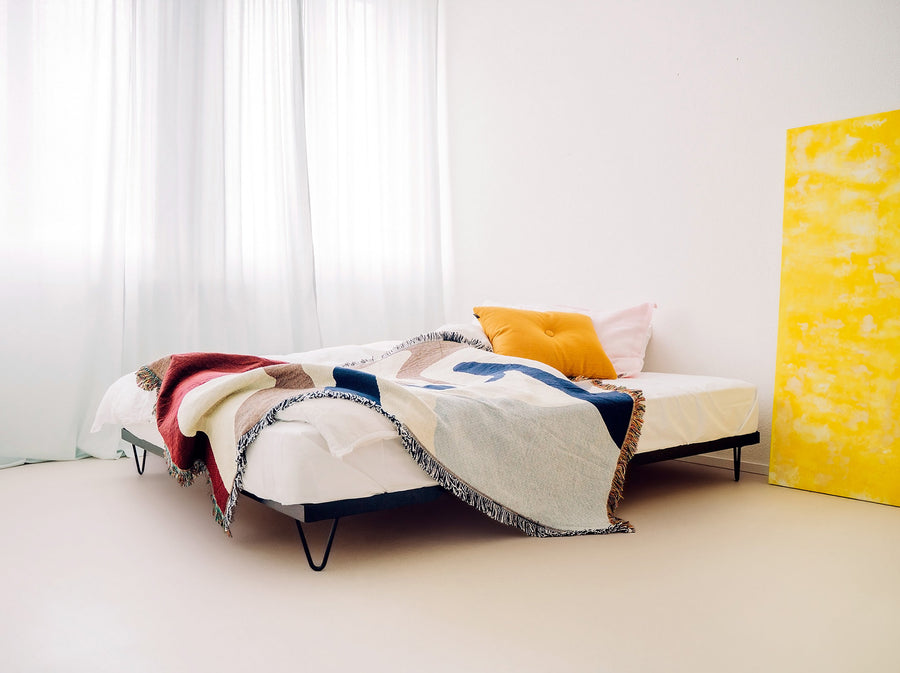 pan mobili, Seitenansicht Kitanda Bettgestell mit weißem Bettlaken, bunter Wolldecke und Kissen darauf in einem hellen Raum mit weißen Vorhängen. 